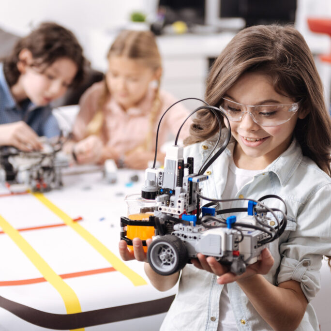 Lovely little girl holding robot at school Smart generation exploring science. Sincere smiling emotional girl standing at school and holding electronic robot while her colleagues working on the project